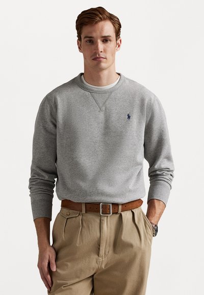 Sweatshirt cinza com punhos e decote canelados, apresentando um pequeno logo azul marinho no peito. Combinado com calças beijas plissadas e um cinto castanho.