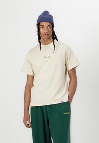 Beige bomulls t-skjorte med grønn grafisk trykk, kortermet, sammen med grønne joggebukser med en subtil logo. Blå stripete lue på modellen.