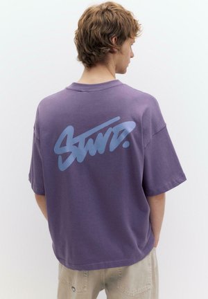 Ung mand med lyst brunt hår iført en løs lilla T-shirt med stor blå stiliseret tekst på ryggen og beige bukser.