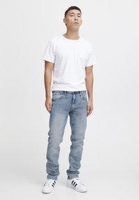 Camiseta blanca de cuello redondo, jeans de mezclilla azul claro y zapatillas blancas con detalles en negro; modelo de pie contra un fondo liso.