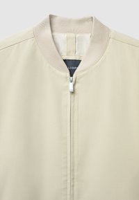 Giacca bomber beige chiaro con colletto a coste, tessuto liscio, chiusura frontale a zip e una leggera lucentezza. Design semplice senza motivi visibili.