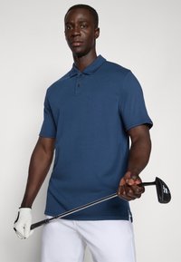 Námořnická modrá polokošile s krátkým rukávem, třemi knoflíky a bočními rozparky, kterou má na sobě osoba držící golfovou hůl, doplněná bílými kalhotami.