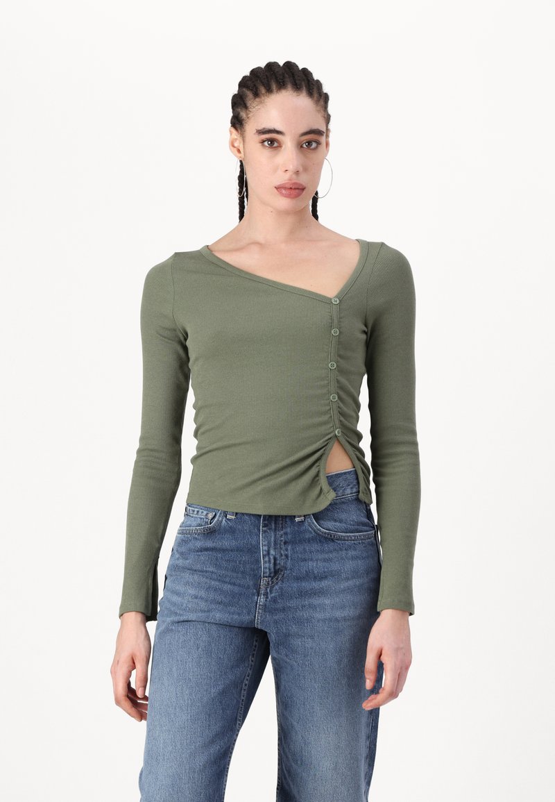 Olivgrön långärmad topp med en v-wrap-design, ribbad textur, knappdetaljering och en sidofrunns accent. Kombinerad med ljusblå jeans.