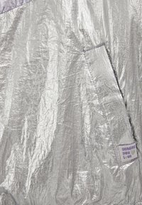 Srebrn bleščeč material z zgubanimi teksturami. Vsebuje vijolično etiketo z napisom »pošiljam ti ljubezen« in detajl žepka.