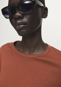 Rjava srajca s kratkimi rokavi in črnimi sončnimi očali. Srajca ima okrogel izrez, sončna očala pa debel okvir in temna stekla.