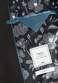 Interno di giacca su misura con motivo floreale in tonalità di grigio, con una tasca accentuata color teal e etichette con scritto "next", "40R" e "SLIM FIT".