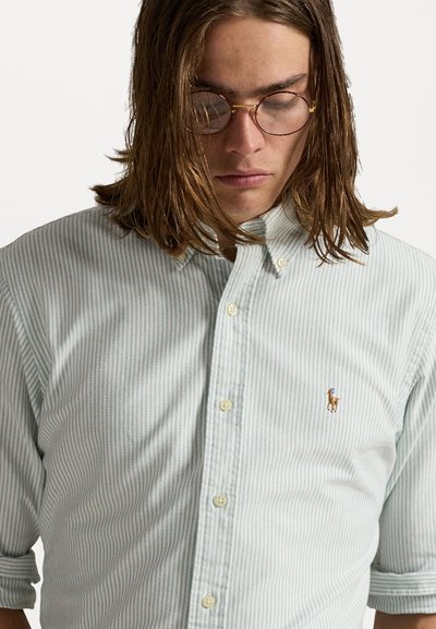 Chemise à rayures bleues claires et blanches avec un col, comportant un logo brodé sur la poitrine. Matière en coton, coupe décontractée.