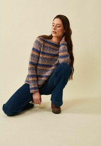 Kvinna som knäböjer med handen på nacken, iklädd en mönstrad tröja i brunt, blått och beige, mörkblå jeans och bruna skor mot en beige bakgrund.