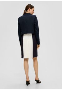 Marineblauw cropped jack over een jurk met een wit middenpaneel, omringd door donkere panelen. Het model draagt zwarte haksandalen.