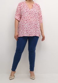 Person iført en bluse med pink og hvid leopardprint, mørkeblå slim-fit jeans og lysebeige spidse sko, stående foran en ensfarvet væg.