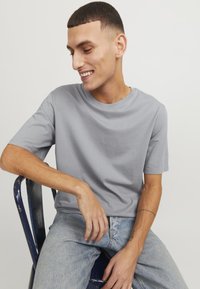 Grijze katoenen T-shirt met een ronde halslijn en korte mouwen, gedragen met lichtblauwe spijkerbroek, zittend op een blauwe metalen stoel.