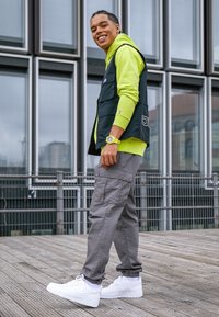 Černá vesta přes neonově zelenou mikinu s kapucí, šedé cargo kalhoty a bílé tenisky. Má strukturovanou tkaninu a viditelné švy.