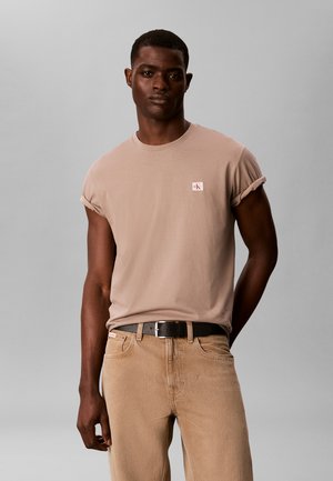 Junger Mann trägt ein hellbraunes T-Shirt mit aufgekrempelten Ärmeln und kleinem Calvin Klein-Logo sowie beige Hosen mit schwarzem Gürtel.