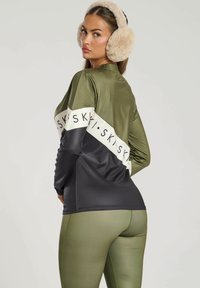 Oliven grøn og sort atletisk top med hvidt bånd; har tekst; parret med oliven grøn leggings. Fuzzy ørevarmere fuldender looket.