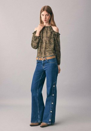 Giovane donna che indossa una blusa leopardata a spalle scoperte, jeans a vita alta e gamba larga con bottoni laterali, cintura marrone e scarpe a punta marroni, in piedi.