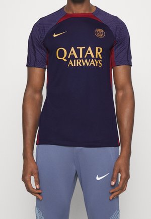 Mężczyzna w granatowo-czerwonej koszulce piłkarskiej Paris Saint-Germain z złotym logo Qatar Airways oraz fioletowych spodniach Nike z białym logo Swoosh.