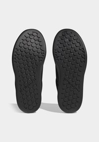 Černé gumové podrážky bot s kruhovým vzorem pro lepší přilnavost, s logem na spodní části. Plný, široký design pro stabilitu a trakci.