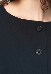 Γυναίκα που φοράει ρούχο με τετράγωνο λαιμόκοψη σε μαύρο χρώμα, με δύο μεγάλα μαύρα κουμπιά και καστανά σγουρά μαλλιά που φαίνονται εν μέρει.