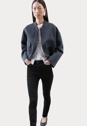 Bomber jacket in lana grigia scura con due tasche frontali, abbinato a una camicia grigia chiara e jeans skinny neri. Modello che indossa scarpe basse.