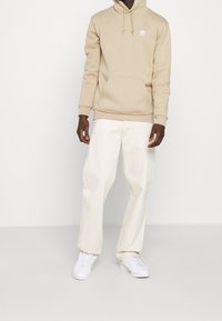 Beige hoodie med framficka och dragsko, tillsammans med krämfärgade vida byxor och vita sneakers, stående mot en neutral bakgrund.