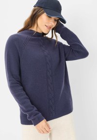 Marineblauwe gebreide sweater met een hoge kraag en kabelpatroon aan de voorkant. Zachte textuur, lange mouwen en geribde manchetten. Caps gedragen.