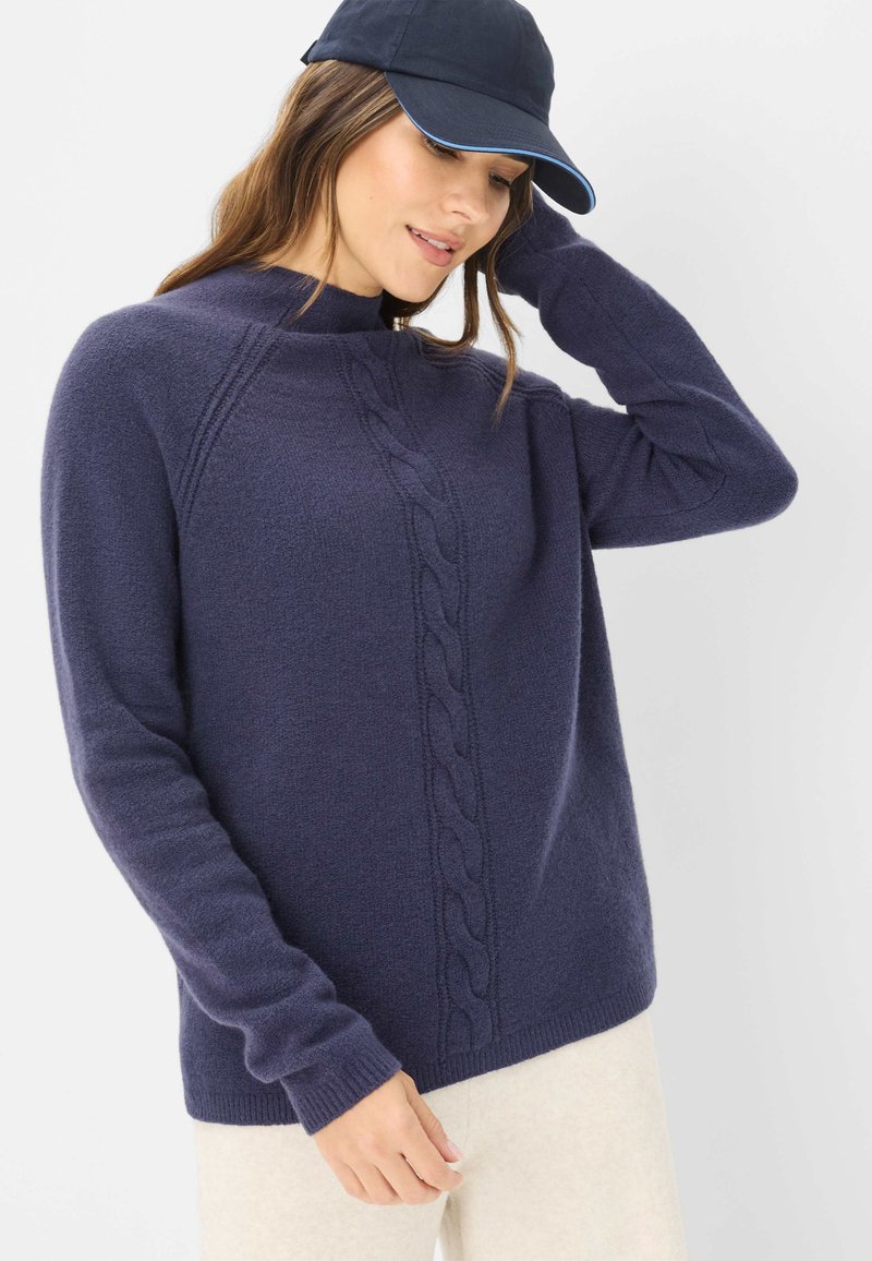 Marineblauer Strickpullover mit hohem Kragen und Zopfmuster auf der Vorderseite. Weiches Material, lange Ärmel und gerippte Bündchen. Mützen getragen.