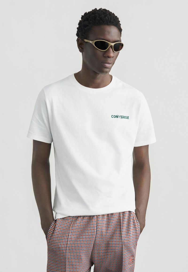 Άσπρο βαμβακερό μπλουζάκι με πράσινο λογότυπο «CONVERSE», φορεμένο με καρό σορτς που έχουν κόκκινα και μπλε σχέδια και πλαϊνές τσέπες. Οι φακοί είναι οβάλ σχήματος.