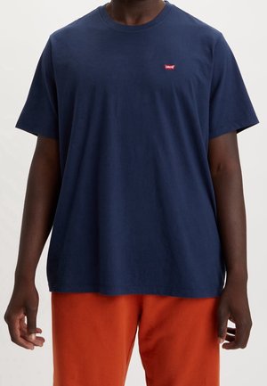 Mężczyzna w granatowej koszulce Levi's i pomarańczowych spodniach, stojący z ramionami swobodnie opuszczonymi wzdłuż ciała na jednolitym tle.