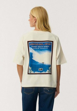 Blond vrouw met de rug naar de camera, draagt een wit T-shirt met een blauwe surfprint en tekst over de Ripcurl Cup op Sunset Beach Hawaii, gecombineerd met een blauwe spijkerbroek.