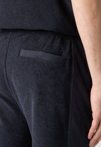 Gros plan sur une personne portant un pantalon texturé foncé avec une poche arrière horizontale et une partie de l'avant-bras nu visible.