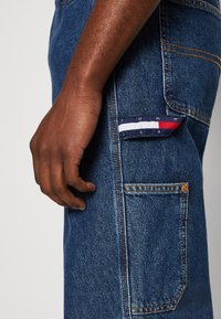 Jeans i denim med en blå tvätt, lätt slitage och en synlig ficka med en tygetikett i röd, vit och blå rand.