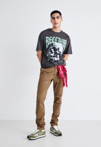 Camiseta gris con gráfico de calavera verde, pantalones beige y zapatillas verde oliva. Bandana roja atada a la cintura. Diseño ajustado y casual.
