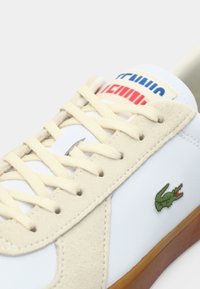 Baskets blanches avec accents en daim beige, présentant des lacets crème, un logo crocodile vert, et une étiquette bleue et rouge sur la languette. Semelle en caoutchouc.