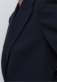 Blazer navy con un diseño estructurado, que presenta solapas afiladas y una silueta ajustada; tejido suave con detalles de costura mínimos.