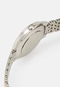 Sølv rustfrit stål ur med rundt urkasse, minimalistisk design, metalarmbånd med cylindrisk led og en stenbesat krone.