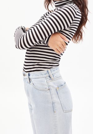 Kobieta z długimi włosami, krzyżująca ramiona, ubrana w czarno-biały, paskowany golf i jasnoniebieskie, wysokoprofilowane dżinsy, ukazana z profilu.