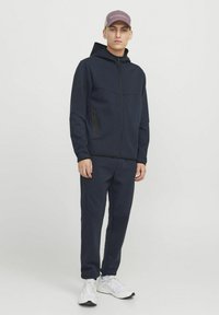 Un sweat à capuche bleu marine avec une fermeture éclair et des poches latérales, associé à un pantalon tapered assorti. Il est fabriqué en tissu lisse et est porté avec des baskets blanches.