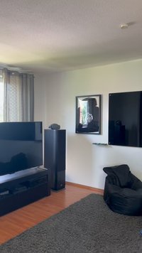Televisor plano preto sobre uma unidade baixa preta, ao lado de um alto altifalante preto. Cadeira de saco preta sobre um tapete cinza texturizado contra uma parede clara.