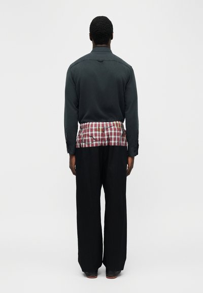 Άνδρας που στέκεται με την πλάτη, φοράει μαύρο πουκάμισο, μαύρο παντελόνι και ένα κόκκινο-άσπρο καρό πουκάμισο δεμένο γύρω από τη μέση.