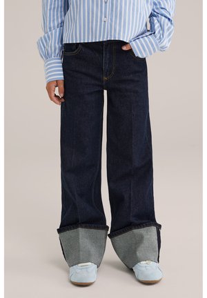 Jeans a gamba larga blu scuro con risvolti in denim grigio, decorati da cuciture gialle e tasche laterali, abbinati a scarpe azzurre.