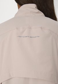 Veste de sport rose clair avec un col haut, une texture lisse et des détails de logo subtils en gris dans le haut du dos.