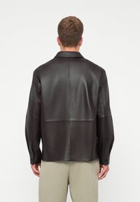 Fekete bőr ingdzseki sima textúrával, hegyes gallérral, hosszú ujjaival és gombos mandzsettákkal. A dizájn panelvarrást tartalmaz.