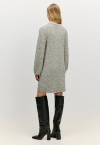 Strickkleid in Hellgrau mit strukturiertem Muster, mit langen Ärmeln und lockerer Passform. Kombiniert mit schwarzen kniehohen Stiefeln mit Absatz.