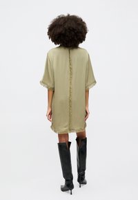 Olivovo zelené šaty s strapcami pozdĺž okrajov. Uvoľnený strih, krátke rukávy, dĺžka po stredný steh. Nosené s čiernymi čižmami do kolien.