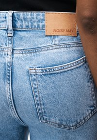 Šviesiai mėlynos spalvos džinsai iš džinsinio audinio, matomas kišenės nugarėlė ir rudos odos prekės ženklo pleistras su užrašu „NOISY MAY“, dėvimi tamsios odos žmogaus.