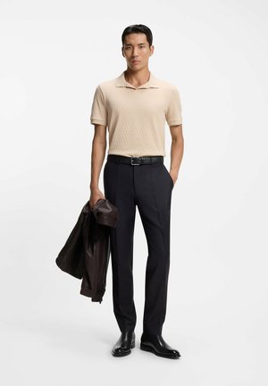 Beige polo shirt met textuurpatroon, donkere op maat gemaakte broek, zwarte riem en zwarte leren schoenen; jas over de arm gedrapeerd.