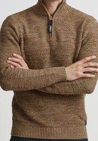 Pull marron en maille avec un col montant et une fermeture éclair à moitié. La texture est structurée, avec des poignets et un ourlet côtelés. Coupe légèrement cintrée.