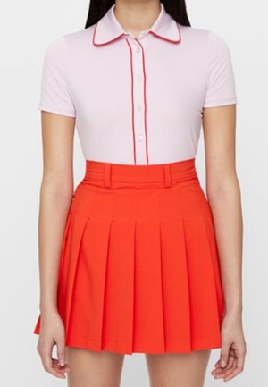 Vrouw die een lichtroze polo met korte mouwen, rood beleg en een hooggetailleerde, felrode geplooide rok draagt.