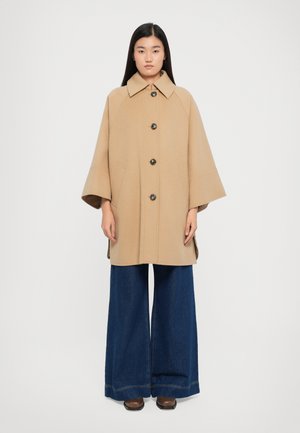 Beige wollen jas met een ruime kraag en knopen aan de voorkant, voorzien van zijzakken en een losse pasvorm, gecombineerd met marineblauwe wijde pijpen jeans.