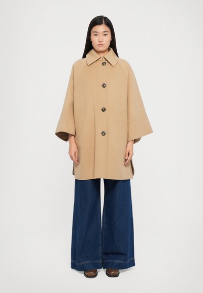 Beige wollen jas met een ruime kraag en knopen aan de voorkant, voorzien van zijzakken en een losse pasvorm, gecombineerd met marineblauwe wijde pijpen jeans.